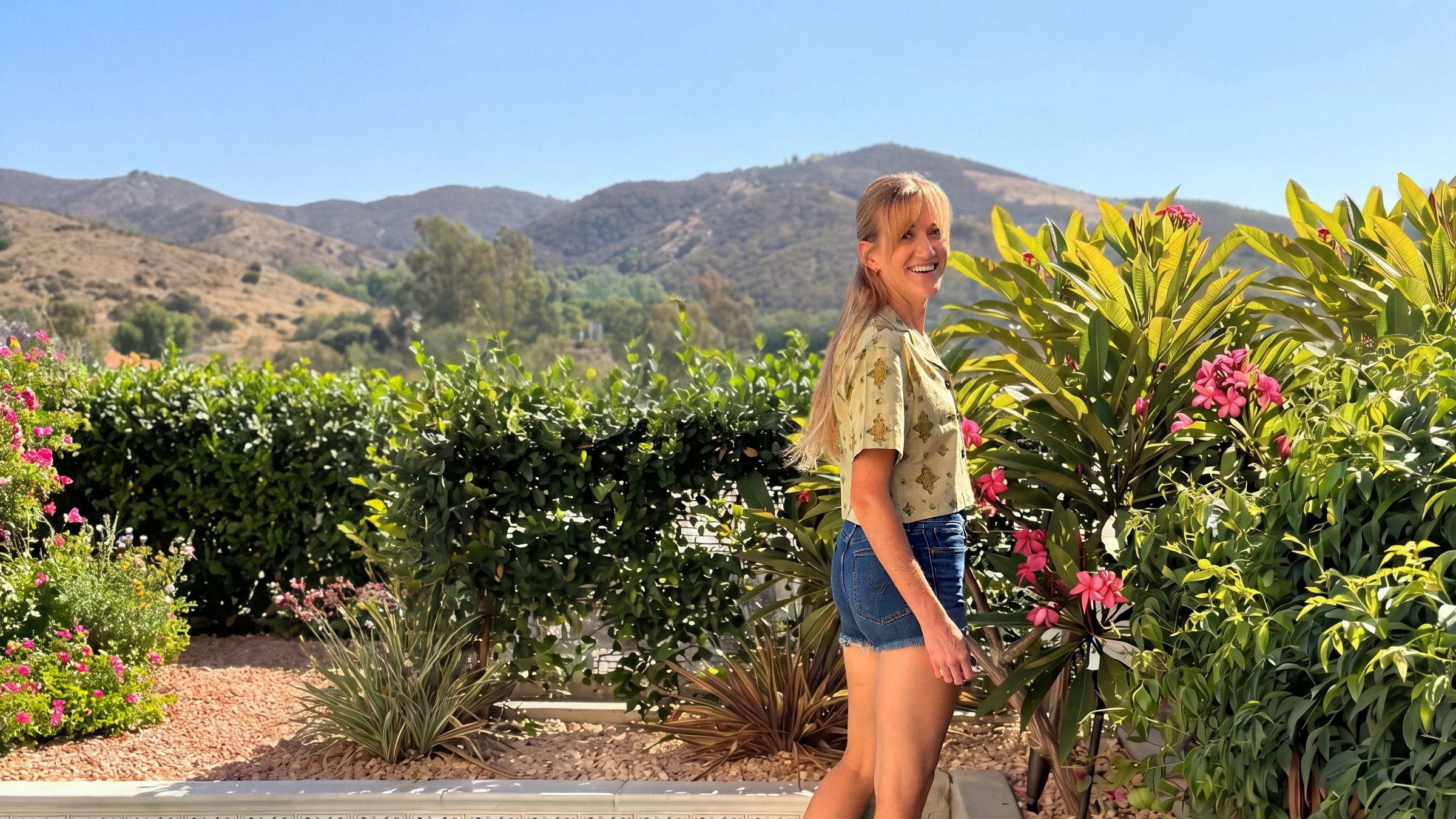 A woman smiling in a garden of plumeria, with hills behind her, wearing a pale yellow block-printed shirt