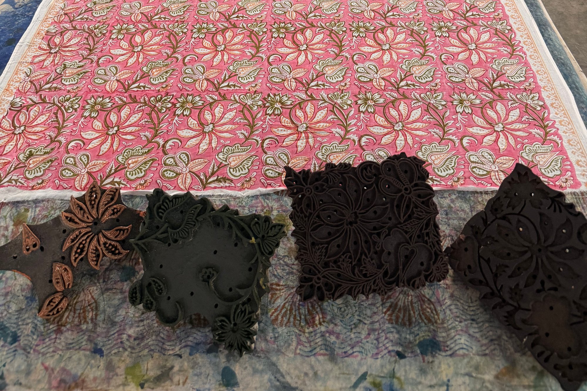 Cotton printed with pink flowers on a printing table, with four carved wooden printing blocks laid in front of it