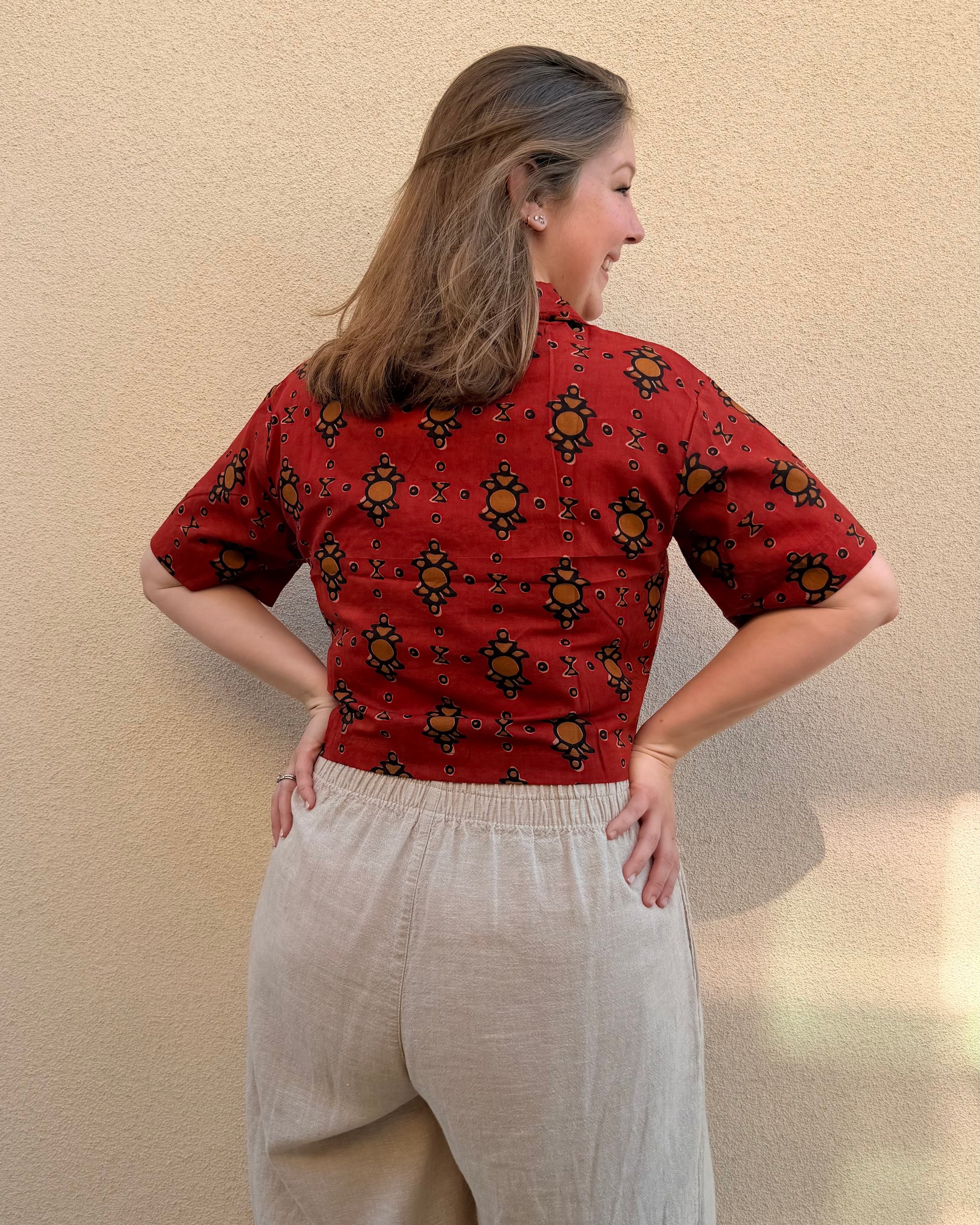 A woman in a red block-printed shirt and linen trousers, seen from behind, smiling over her shoulder
