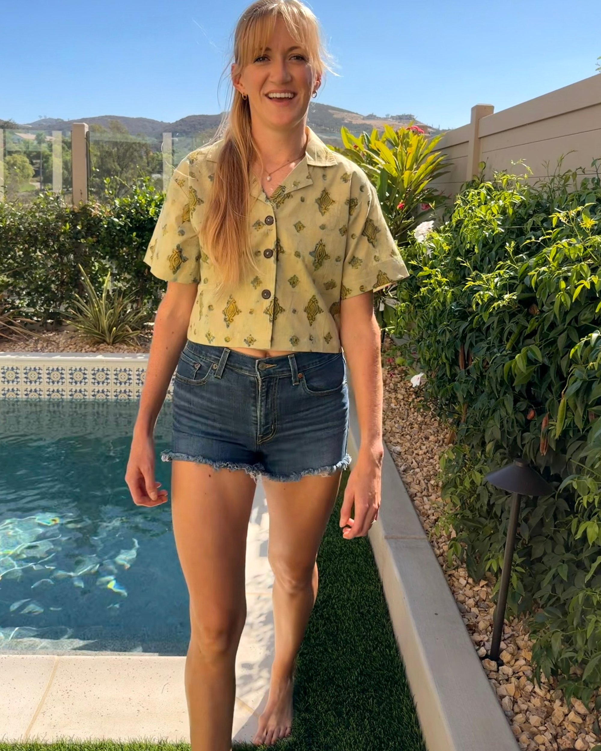 A woman walking beside a pool in a pale yellow block-printed top, with hills behind her